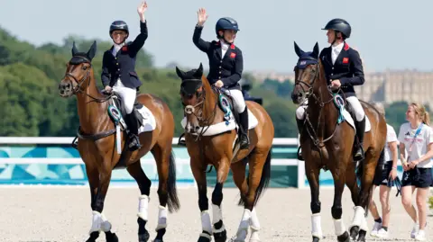 EPA Ros Canter, Laura Collett and Tom McEwen wave to the crowds as they arrive at the medal ceremony on their horses, which are wearing rosettes.