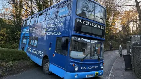 A blue double decker bus with a number along the top and the words "Eurocoaches, Eurominibuses, Eurotaxis" on the side. Its front end is blocking a narrow road, which bends to the right behind it. The back of the bus appears to have reversed over a wall/hedge off to the side of the road and is tilted upwards.