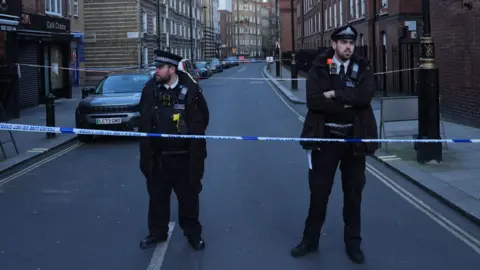 PA Media Two police officers standing behind police tape. Both are men dressed in black uniforms. Behind them is a street with high brick buildings either side and parked cars