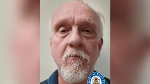A mugshot of a man with white hair and a white beard. He looks at the camera. A West Midlands Police logo is at the bottom of the photo.