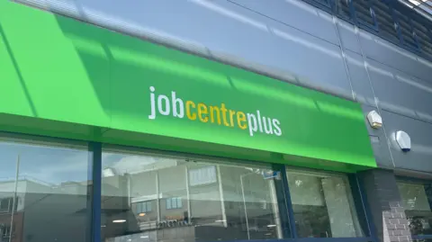 The Jobcentre Plus sign, with white and yellow lettering on a green background, with a window underneath reflecting the street opposite