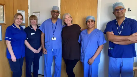 Five people in nurse outfits or scrubs standing with a woman wearing an all black outfit with shoulder-length white hair.