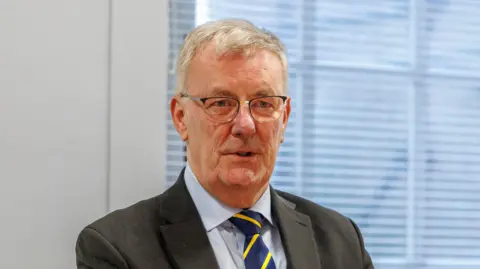 Liam McBurney/PA Wire Mike Nesbitt, a man with short, grey hair and glasses, at an event in Stormont last month.  He is wearing a black suit, a blue shirt and a navy tie with a yellow stripe. He is standing in front of a window in a white room.