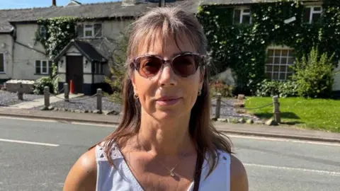 There is a woman with brown shoulder length hair, and a fringe. She is wearing brown-tinted sunglasses and two hoop earrings. She has a white sleeveless vest. Behind her is the closed down pub and all of the windows have been covered by blinds.