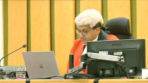 A judge in a red robe and white wig looking at a laptop in a courtroom with wooden panelling