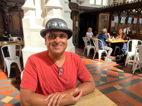 A man sits in a church cafe wearing a red shirt and a black hat with sunglasses on top. 