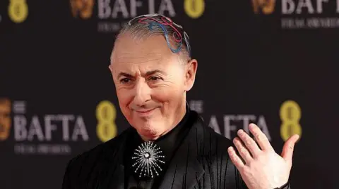 Getty Images Alan Cumming with colourful coils on his head and a silver, diamond brooch and black jacket and shirt