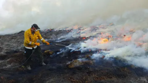 RSPB Scotland A firefighter wearing protective clothing uses a hose to spray water on a smouldering moorland fire. Flames and thick white smoke spread across the burnt vegetation in the background as firefighting efforts continue.