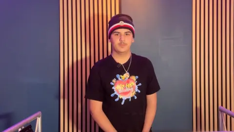A picture of Sam standing up in front of a dark blue wall with vertically panelled wooden slats. He wears a black red and white hat with a black shirt with a colourful Hip-Hop Healing graphic.