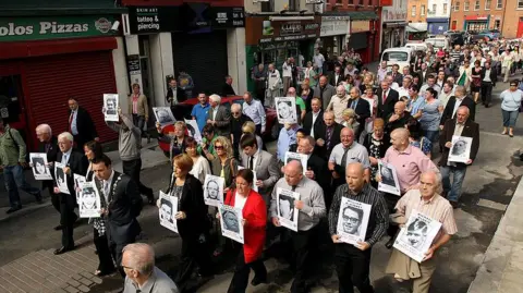 Getty Images Families of the victims of Bloody Sunday carrying black and white images of their relatives, which is on white paper. They are dressed in a variety of coloured clothing as they walk past shops and restaurants on the streets of Londonderry.