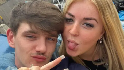 Tyler Kerry and his girlfriend pose in a selfie, with the girlfriend sticking her tongue out. He has short brown hair and she has long blonde hair.