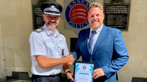 Essex Police Glen Pavelin is wearing his police uniform and is shaking Singer's hand. Singer is dressed in a blue shirt and navy tie and is holding the certificate as the award is presented.