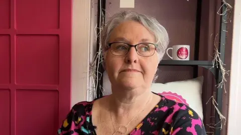 Lindsey Brown, who has short grey hair, sitting in front of a black, metal book case with fairy lights wound round it. She's wearing black glasses,  a top with a bright neon splatter pattern on it and a silver necklace.