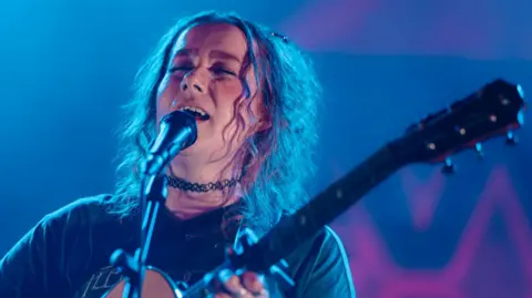 Nati has her eyes closed as she sings into a microphone and plays a guitar. She has curly hair down to her shoulders. The stage inside the festival's big top is blue and pink.