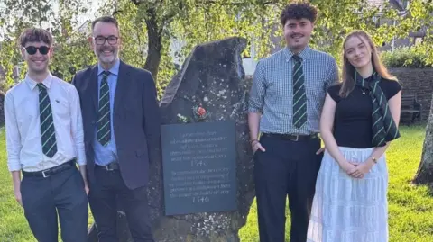 Caleb Tutt Four people smiling at the camera next to a big rock with an inscribed plaque on it. Three of the people are men, and one is a woman. Far left a man has dark sunglasses on, a white shirt, a green, blue and purple tie and blue trousers, next to him is a man with a salt and pepper goatee, blue blazer and blue trousers with a light blue shirt and the same coloured tie. The rock is in the middle, next to the rock (on the right) is a man with curly brown hair, a white, green and purple checked shirt, with blue trousers and the same coloured tie. And next to him is a woman with shoulder-length brown/blonde hair with a blue t-shirt and white skirt with a striped necktie in the same colours as the men's ties.