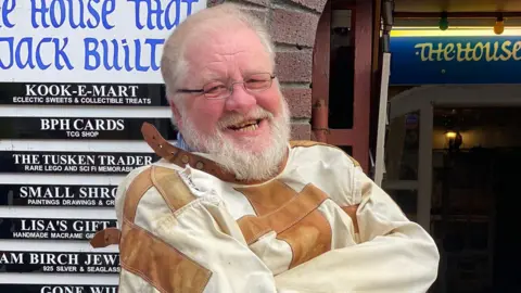 Malcolm Norton who has a white beard, glasses on and a broad toothy smile. He is wearing the white and brown straitjacket that has been returned to his shop. Malcolm is stood in front of his shop's entrance.