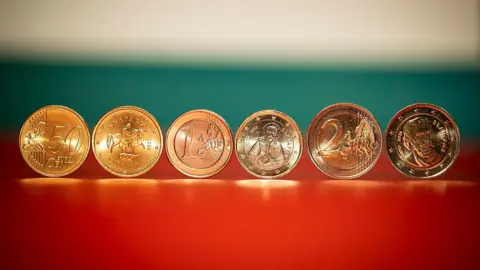 Nikolay DOYCHINOV / AFP via Getty Images Close up of six gold coins showing the symbols on them, including a person on a horse and a face and a saint-type figure
