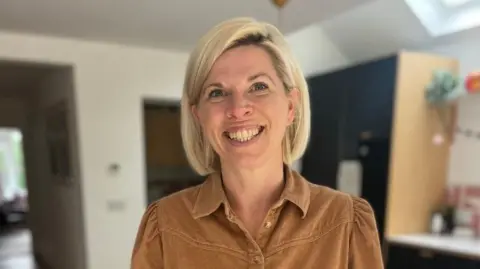 A woman in her late thirties/early forties is standing in her kitchen, smiling at the camera. She has a sandy brown shirt on and has a blonde bob haircut.