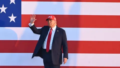 Donald Trump waves wearing a baseball cap and suit in front of a huge US flag poster