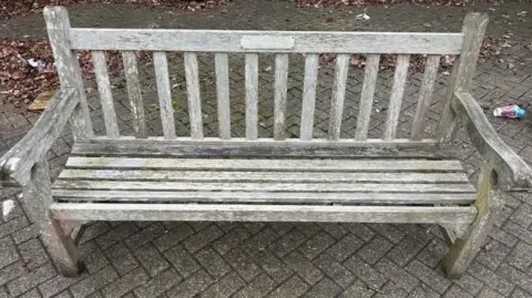 A brown wooden bench that has been bleached by the sun and the elements. A small plaque is top centre of the backrest, the words cannot be made out. The bench sits on grey pavement tiles