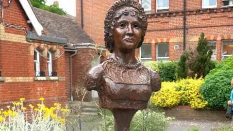 A close-up of the statue, which is a bronze bust of Jane Austen attached to a pile of books. The Regency-style garden can be seen behind it.