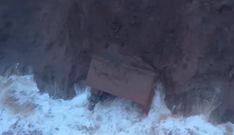 Timothy's Travels Still shot of the bunker at foot of the cliff taken from drone footage. There are waves hitting the structure which appears to be largely intact.