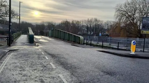 Bristol City Council Image shows an empty road in central Bristol with a bridge with green railings. Behind is a cyclist and trees with no leaves on. 