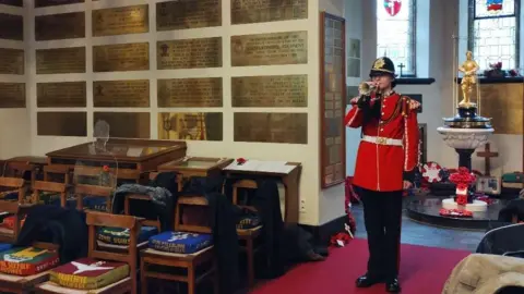 Crown Copyright A soldier stands in a church playing a bugle. He wears a red buttoned up jacket, black hat and black trousers. There is a font behind him with a cross made of poppies at the foot, and a gold statue on top. Brass plaques line the wall.