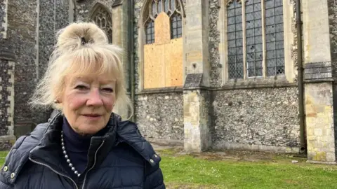 Andrew Turner/BBC A woman standing outside a church, which has one window boarded up and another is damaged. She has blonde air which is tied up and she is wearing a dark anorak, a blue polo neck jersey and a pearl necklace.