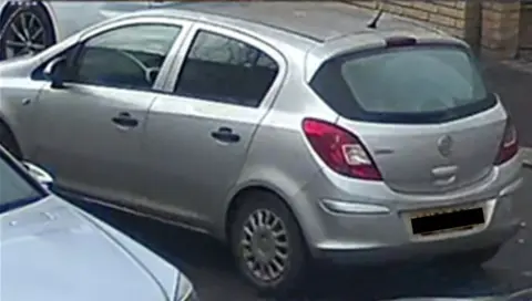 Northamptonshire Police A CCTV photo of a silver Vauxhall Corsa driving down a road past two other parked cars.
