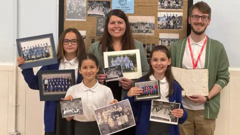 BBC Five people are standing in front of a board displaying old photographs on a wall. Ffion, a pupil has brown hair and black glasses, she is holding two colour photographs of school children. Caia has brown hair tied back and is holding two black and white photos of schoolchildren. Emilee has long brown hair and is holding one black and white school photos and one in colour. Headteacher Hannah Gharu has brown hair and is holding a black and white school photo and a colour school photo. Tom Ollivier has brown hair and glasses, with a green cardigan. He is holding an old log book