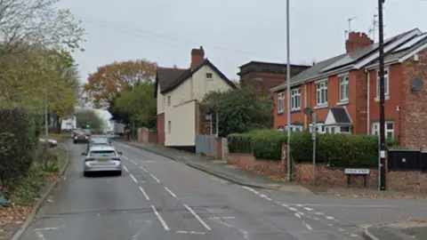 Cars drive up the main road on the left and houses can be seen on the right side of the road before a turning for Fowler Street.