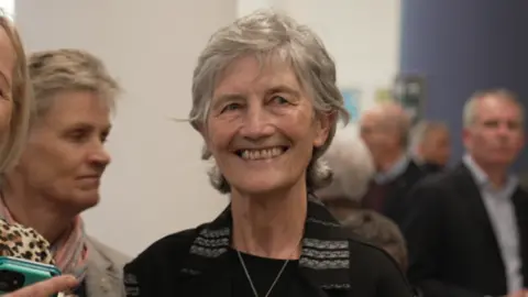 BBC Connolly has short grey hair and is smiling in the picture as she wears a black top and jacket.