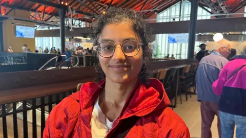 Woman wearing glasses and red coat standing inside market.