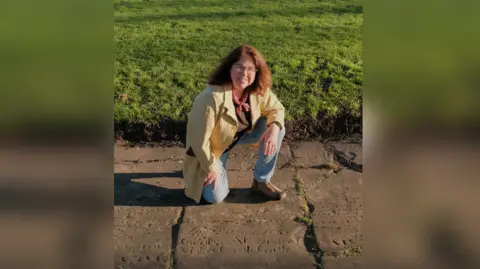 Dr Myra Giesen, a woman with shoulder length brown hair worn down and glasses, kneels on a flat, inscribed gravestone as she smiles at the camera