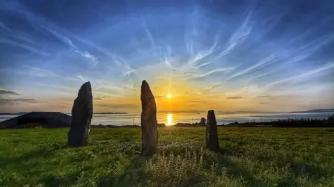 Aileen Gillies Three tall, slender stones standing upright in a grassy field at sunset. The setting sun casts a warm, golden light over the calm sea in the background, which is framed by a low-lying coastline. The sky above is a vibrant mix of blues and oranges, with streaks of cirrus clouds