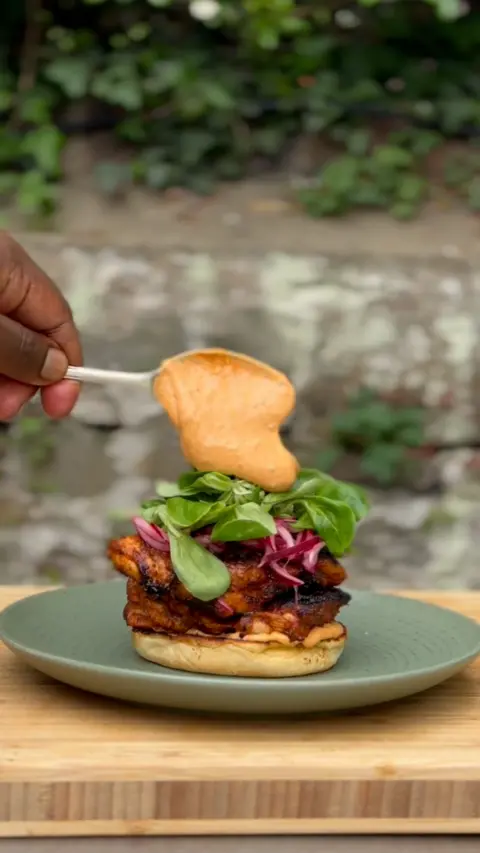 A hand spoons sauce on top of a burger topped with salad