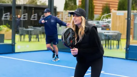 The Padel Club A woman waits at the net on a padel ball court with her racquet raised
