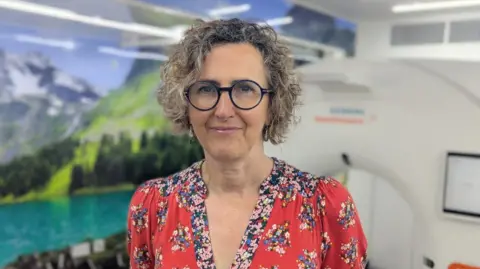 Dr Fiona MacLeod is in the mobile lung cancer screening truck. She has curly hair and round glasses. The white doughnut of the CT scanner can be seen behind her. The wall is decorated with a large photograph of a lake surrounded by mountains.