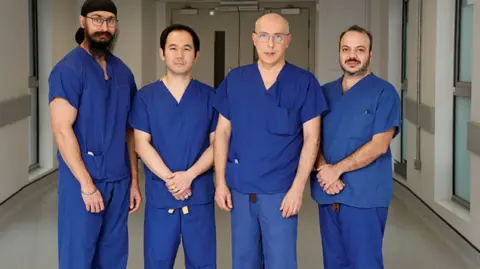 Four men are standing in a line in a hospital corridor wearing blue surgical uniforms. The men are Ranjit Bains, consultant anaesthetist, Dr Lampson Fan, Mr Giuseppe Rescigno and Mr Mahmoud Abdelaziz.