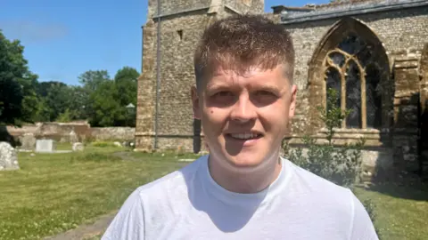 Seth Dellow wearing a white t-shirt standing in the graveyard with the church building behind him 