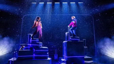 Matthew Murphy Christiani Pitts and Sam Tutty, standing on suitcases on a stage, with lights all around them. It looks like it is snowing on stage. 