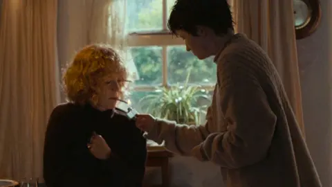 MAB Rabbit Ltd 2024 A curly-haired blond woman sat down, getting her cigarette lit by a short-haired brunette girl holding a lighter. Behind them is a window showing some green trees and sat on the window shelf is a green plant. 