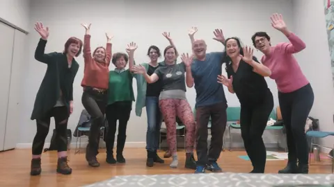 Laughter Yoga with Joe Seven women, alongside Joe Hoare, standing in a line with the hands in the air, smiling