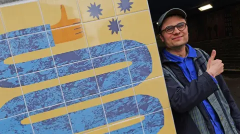 Newcastle City Council Benji Spence, a middle aged man wearing glasses and a cap, giving a thumbs up next to his artwork, which depicts a cartoonishly long arm curving round and round like a snake - also giving a thumbs up.