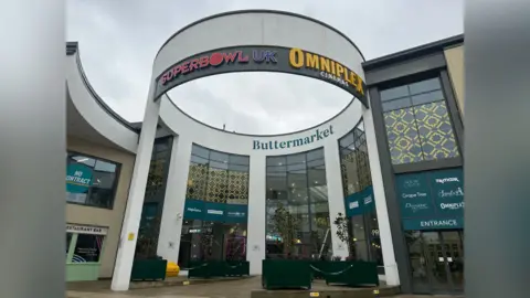 Alice Cunningham/BBC One of the entrances into the Buttermarket shopping centre. The entrance and building is shaped in a semi-circle with a large overhead structure detailing some of the businesses inside the centre