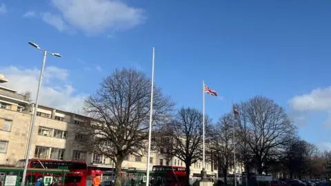 BBC The Royal Parade in Plymouth with flagpoles and buses on the road. One of the flagpoles is flying the Union flag.