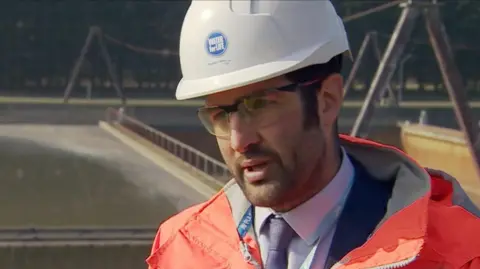 Keith Herbert looks slightly off camera while giving an interview. He has short black hair and stubble. He is wearing protective clothing, a white helmet with a blue circular Southern Water logo, protective clear glasses, an orange high-vis jacket with a white shirt, purple tie and blue blazer underneath. The background is slightly blurred, but shows he is standing in front of Peel Common wastewater treatment works.