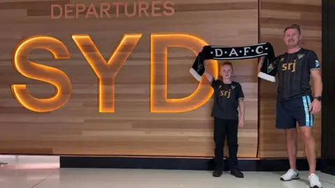 Mark Smith A man and his son in Dunfermline Athletic tops hold up a scarf next to the sign at Sydney Airport