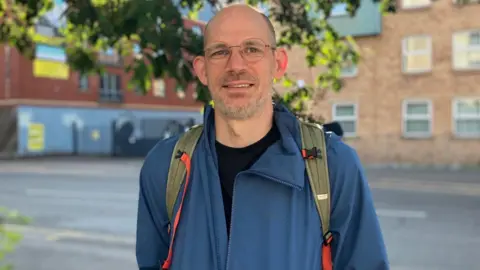 A man in a blue coat and wearing a backpack stands in front of a road. The branches of a tree can be seen in the background as well as several buildings.